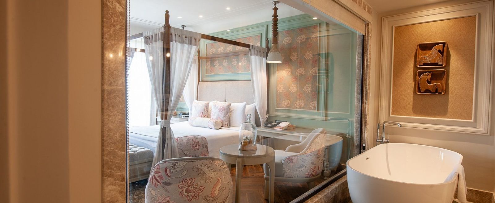A spacious and modern bathroom in a suite at The Evren, Vagator, featuring a freestanding white bathtub. The bathroom is connected to the bedroom by a large glass window, offering a view of the four-poster bed and seating area.