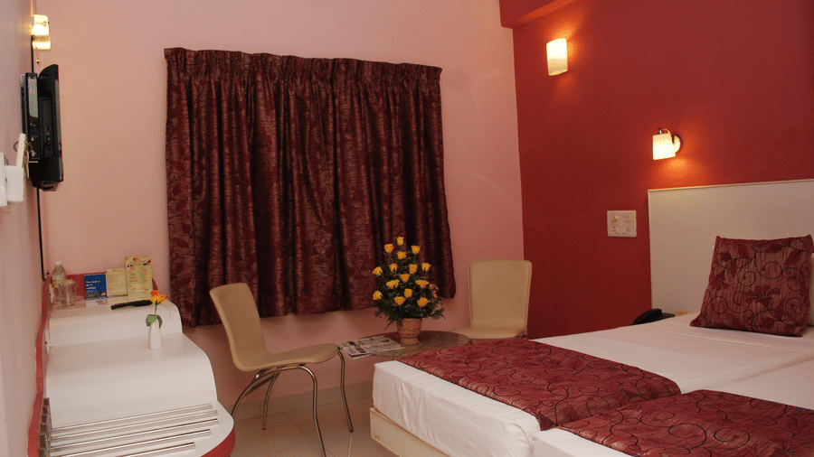 Cosy hotel bedroom featuring adjoined twin beds with maroon throws, light pink walls, and a window with matching dark drapes - Hotel Pandian