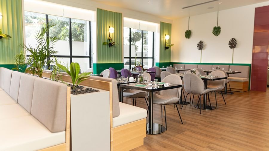 Bright, modern Meli-Melo restaurant interior with accents, large windows, potted plants, and neatly set tables with chairs and beige booth seating. Natural light fills the stylish dining area