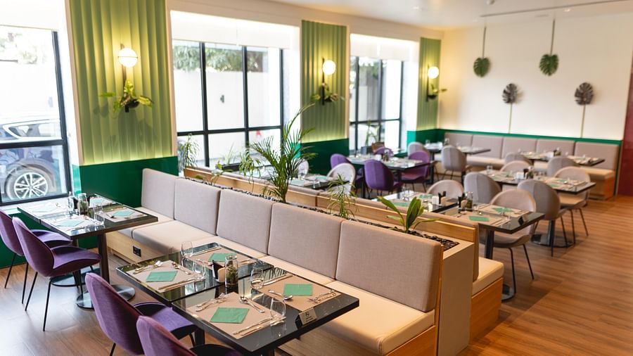 Bright, modern Meli-Melo restaurant interior with accents, large windows, potted plants, and neatly set tables with chairs and beige booth seating. Natural light fills the stylish dining area