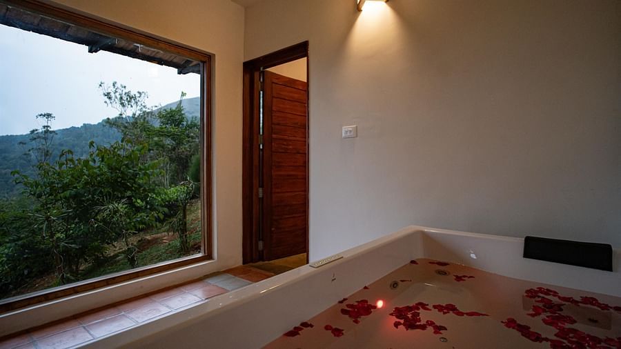 A luxurious bathroom at Vaasta Vagamon with a large tub filled with water and flower petals, beside a window overlooking a natural landscape.