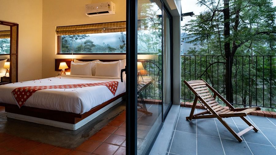 A spacious room at Vaasta Vagamon with a comfortable bed, leading to a private balcony with a wooden lounge chair and scenic views.