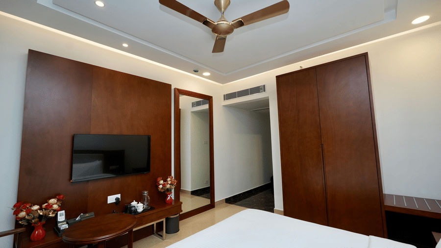 The modern Deluxe King Room at Le Royale Palace with neatly made bed, a ceiling fan, a wall-mounted TV, and wooden furnishing