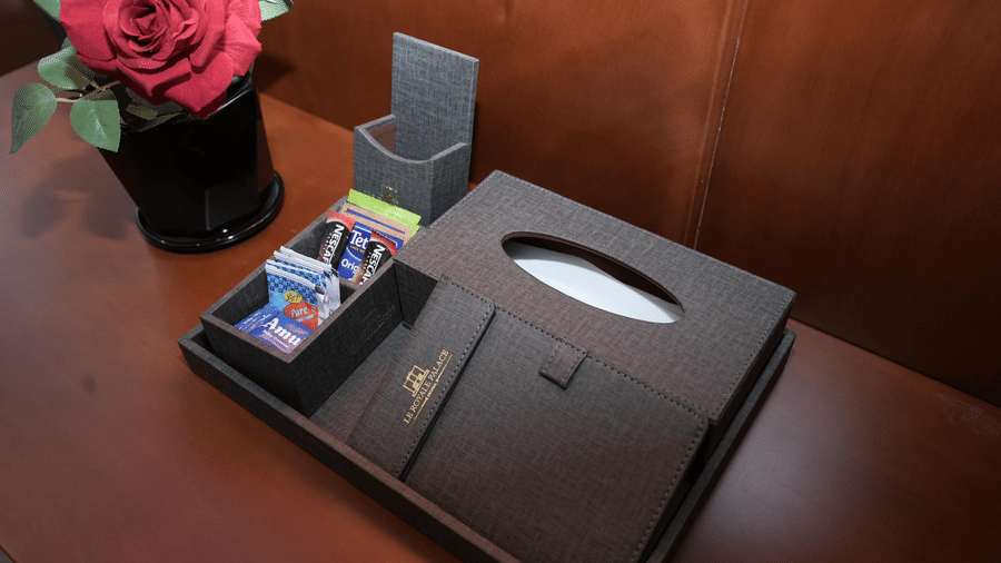 A bedside setup in the Deluxe Quadruple Room at Le Royale Palace featuring a leather stationery box, tissue holder, and a vase with a red rose