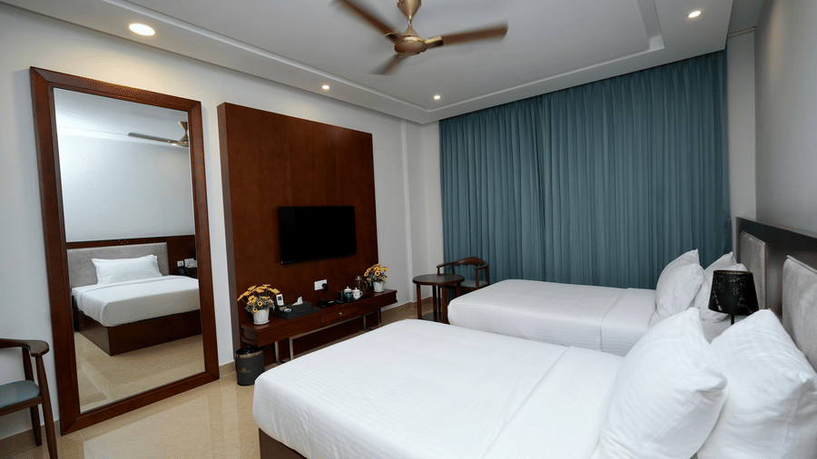 A side view of of Deluxe Twin Room with twin beds, a large mirror, a flat-screen TV mounted to the wall, and a large window with draped curtains