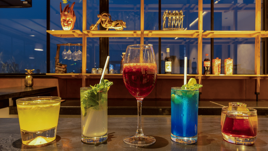 Selection of drinks arranged on a bar counter at Koi Garden. Five distinct glasses contain beverages with ice, straws, and garnishes. Shelving stands behind the counter against a window.