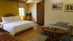 Superior Room at The Richmond, Puducherry featuring a plush bed along with a lamp next to it and a sitting area at the other end of the room with a tiny sofa and a wooden coffee table placed on a carpet