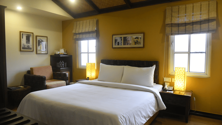 Deluxe Room at The Richmond, Puducherry featuring a plush bed with fresh linen and two lamps placed on the both sides on the nightstand as the sunlight makes its way through the two windows next to a sitting space