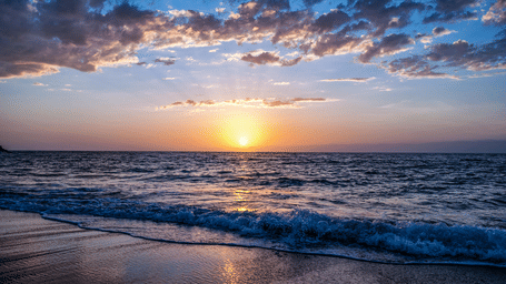 Sun setting over the sea with waves reaching the sandy beach and scattered clouds across the evening sky.