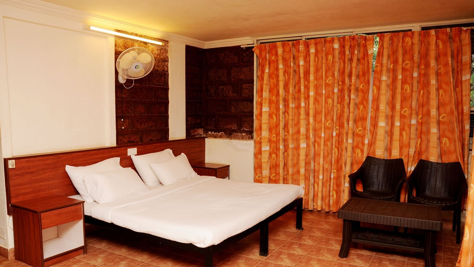 A spacious room at Wind Chalet Resort with a large bed and pillows against a wooden headboard, a bedside table, and a table with a pair of chairs at the foot of the bed, with curtains covering the windows.