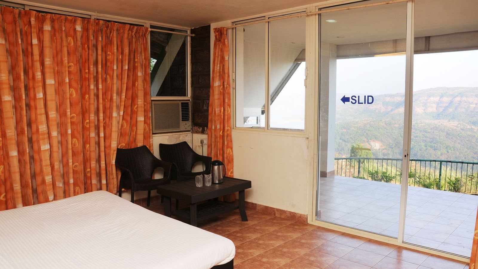 A spacious room at Wind Chalet Resort with curtains covering the windows, a table and chairs placed in the corner, and a balcony offering a view of nature.