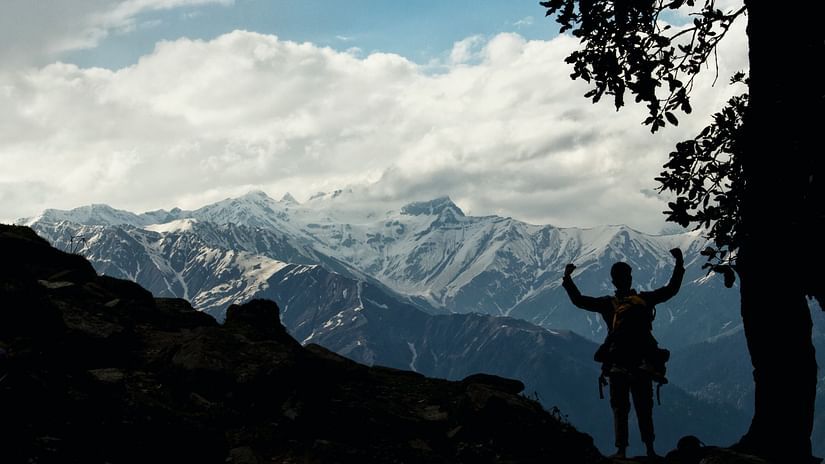 silhouette of a person standing with their arms raised atop a hill with a snow capped mountain range within sight