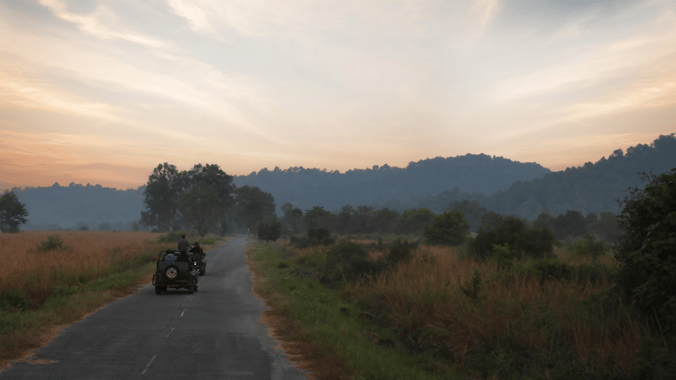 A dirt road leading toward hills at sunset or sunrise, and a vehicle in the distance at The Golden Tusk, Jim Corbett