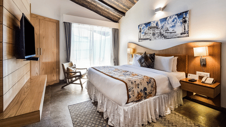 A room with a sloped wooden ceiling, featuring a neatly made bed with a patterned runner and cushions, a wall-mounted television, a wooden wardrobe, and a chair beside a window with curtains, offering a comfortable stay in Udaipur at Bamboo Saa Resort & Sp