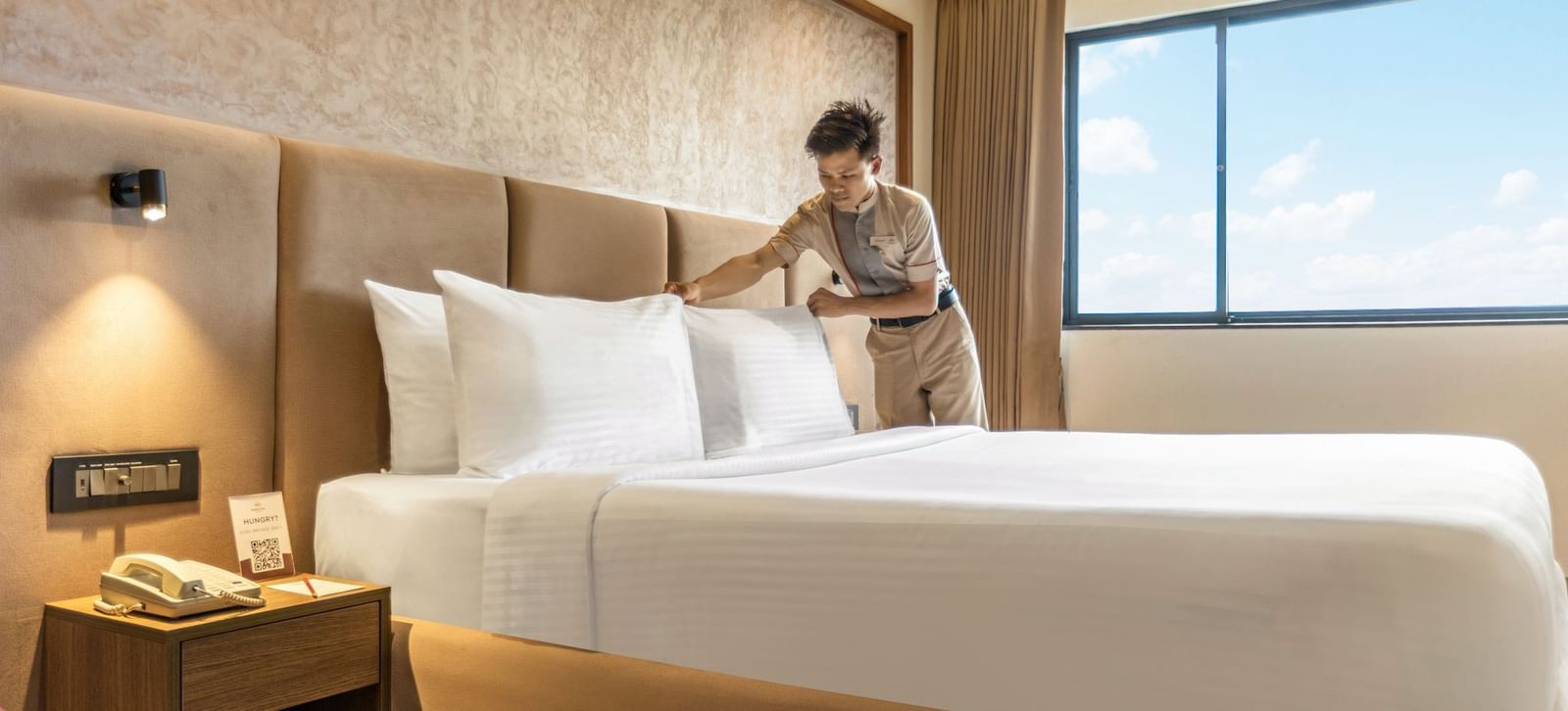 A person arranging pillows on a bed in a room at Mango Hill Central, Chennai. There is a large window on the far wall.