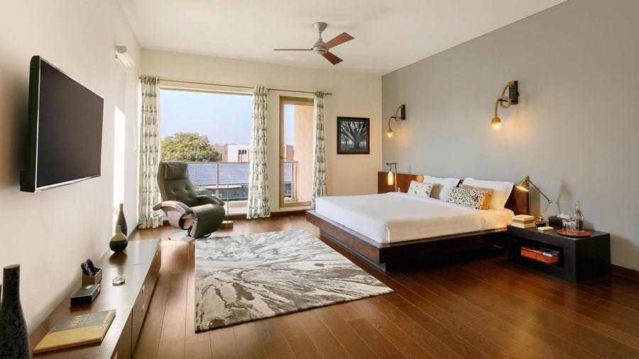 A spacious bedroom with dark wood floors, a low-profile bed, a large window leading to a balcony, a ceiling fan, and a wall-mounted TV - Karma Lakelands