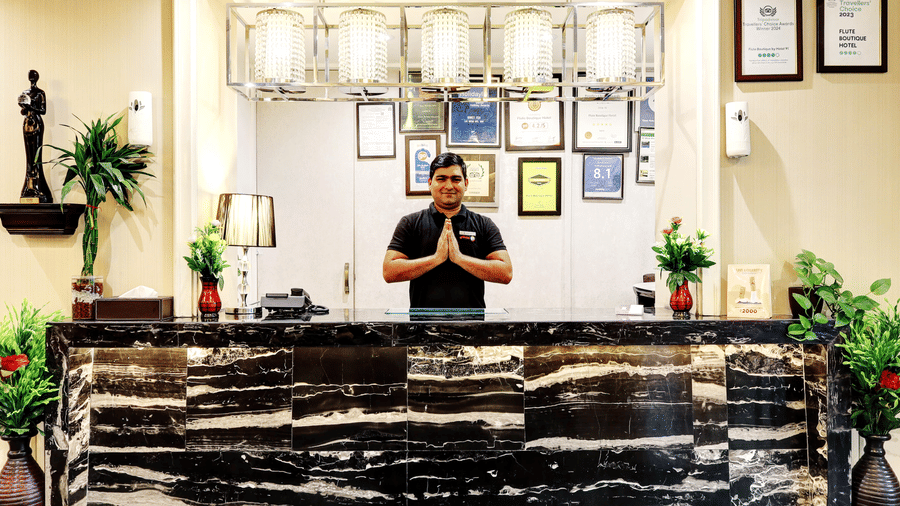 A friendly hotel staff member standing behind the black marble front desk with hands in a traditional Namaste greeting - Flute Boutique By Hotel 91, Jaipur