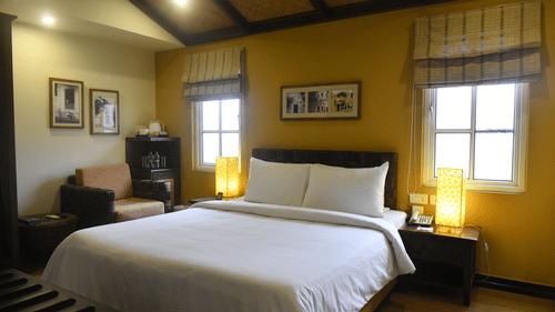 Deluxe Room at The Richmond, Puducherry featuring a plush bed with fresh linen and two lamps placed on the both sides on the nightstand as the sunlight makes its way through the two windows next to a sitting space
