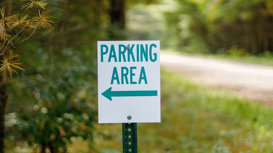White parking area directional sign with a left-pointing arrow displayed on a post beside a green wooded pathway, guiding visitors towards the designated parking area.