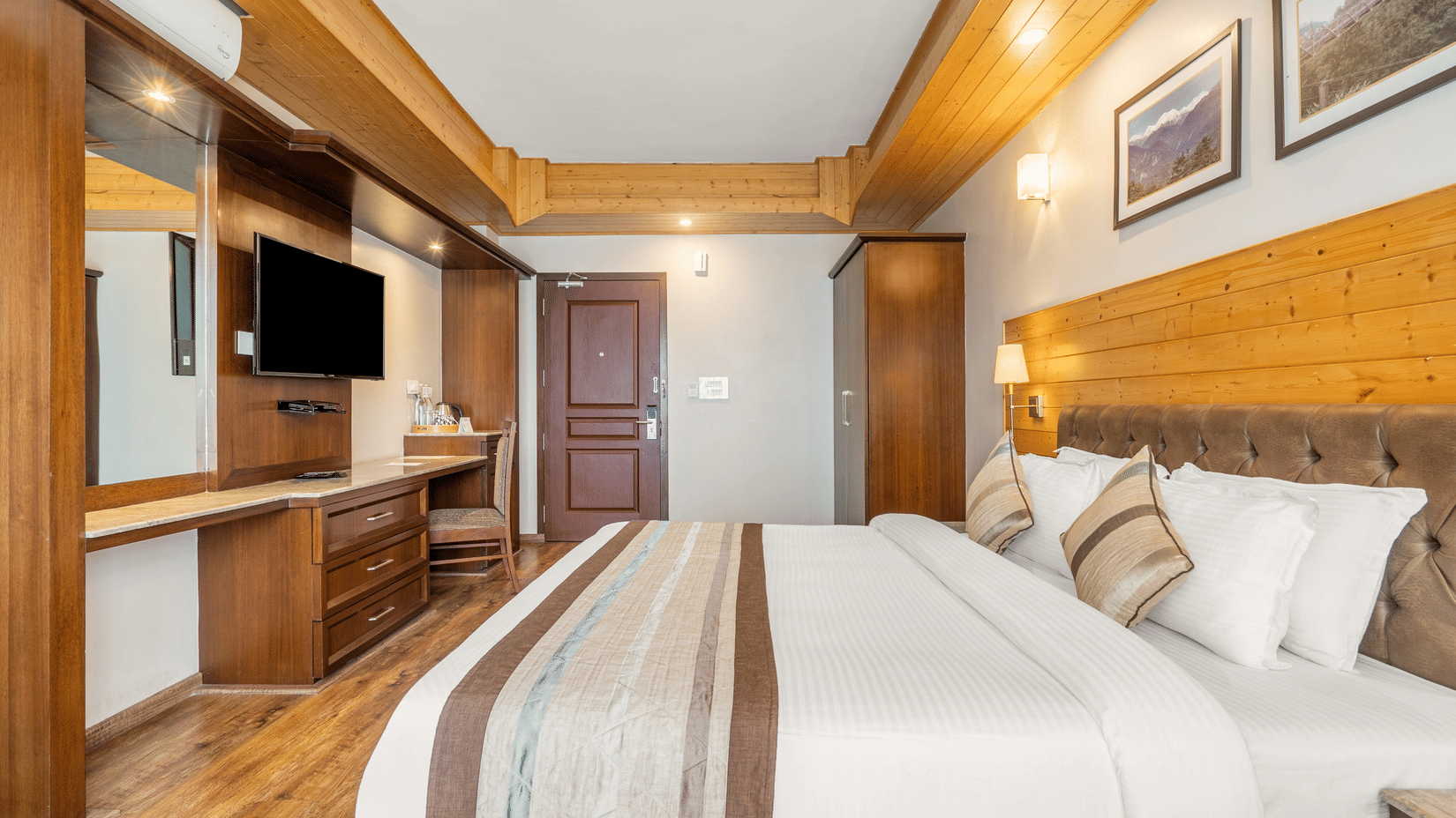 A hotel bedroom featuring a bed with white linens and a striped runner, wooden wall panelling, a television, and a built-in dressing table.