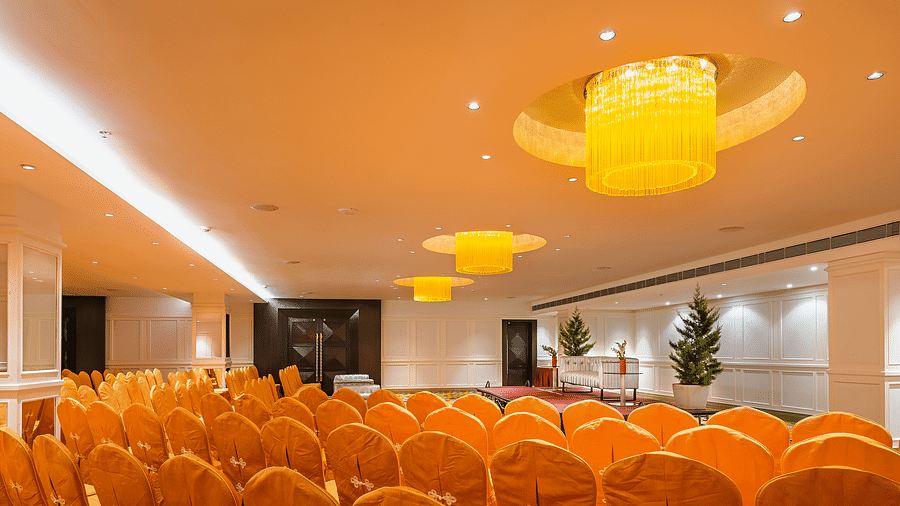 Brightly lit grand ballroom with yellow lighting, white walls, orange seating and a stage with a Christmas tree and red carpet - TGI Grand Fortuna, Hosur