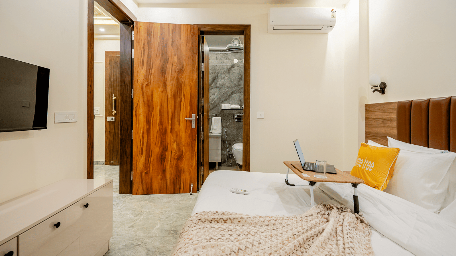 A view inside a hotel room showing a bed equipped with a laptop tray, adjacent to an open wooden door leading into the bathroom.