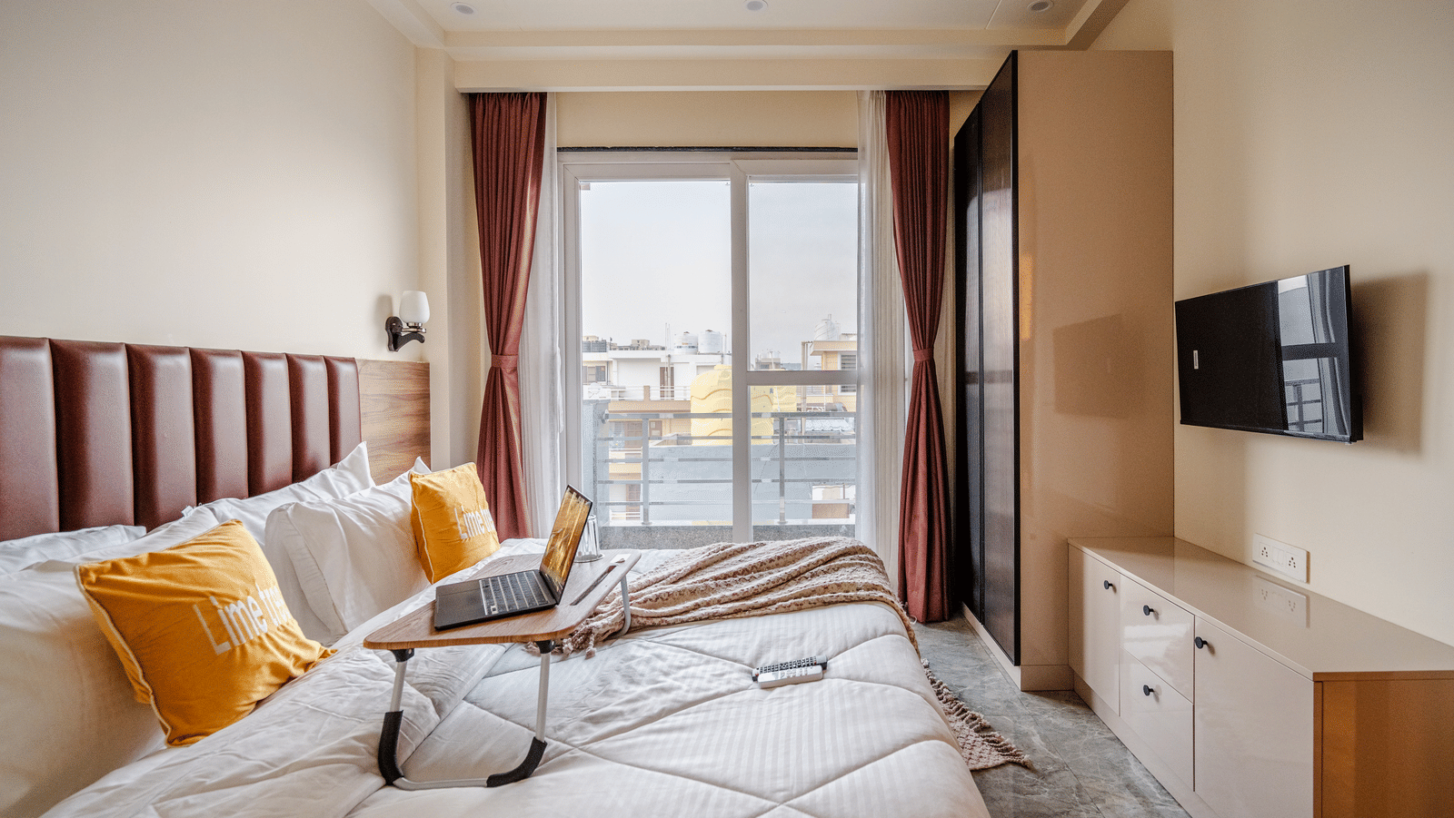 A bright hotel room featuring a bed with a wooden laptop bed tray, yellow accent cushions, and large glass windows.
