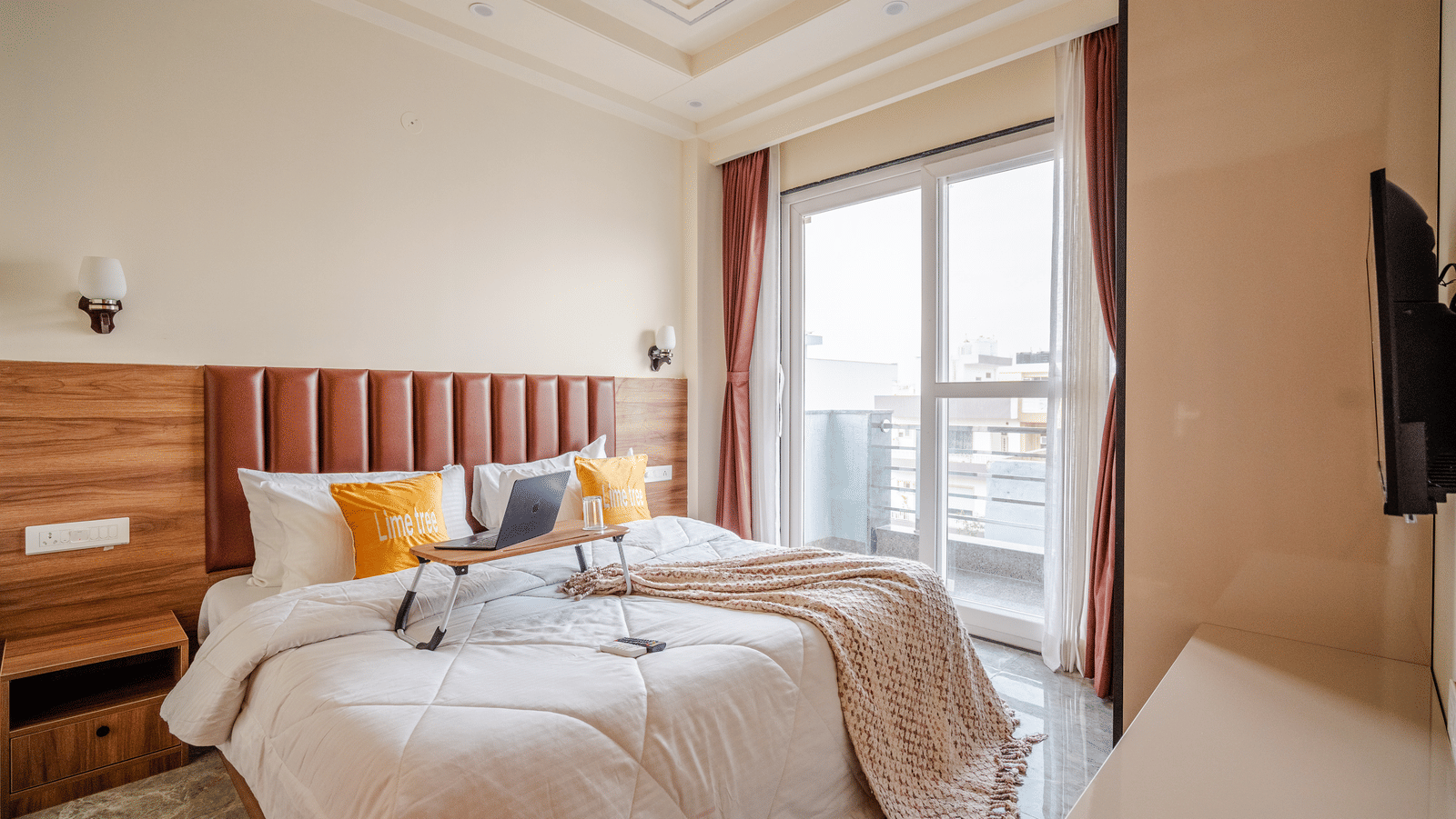 A well-lit hotel room containing a double bed decorated with yellow accent pillows, situated next to large sliding glass balcony doors.