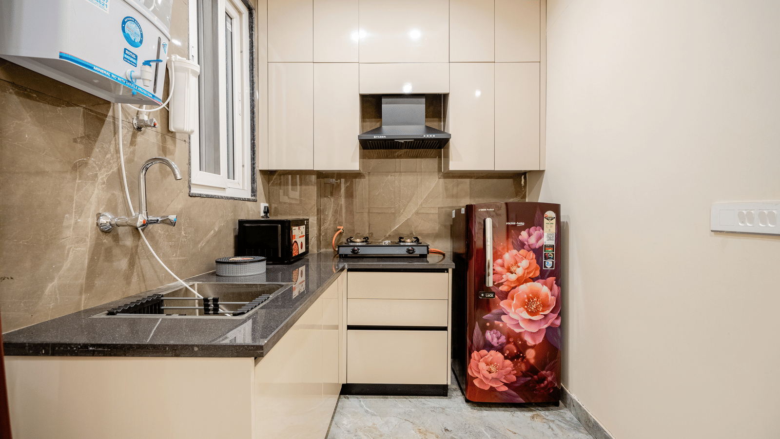 A neat, compact apartment kitchen space showing overhead cabinets, a built-in cooktop stove, a sink, and a floral-patterned fridge.