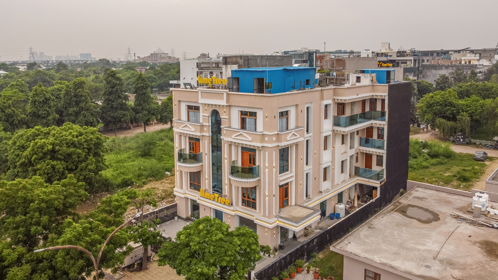 Aerial view of a multiple storey building exterior of Lime Tree Hotels & Service Apartment Private Limited surrounded by trees and neighbouring structures.