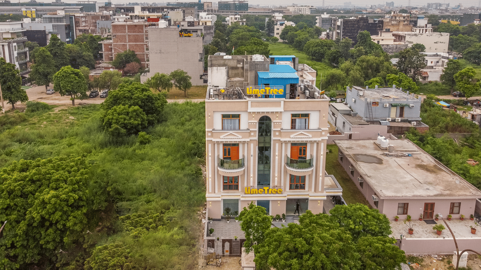 Aerial view of a multiple storey building exterior of Lime Tree Hotels & Service Apartment Private Limited overlooking the surrounding cityscape and greenery.
