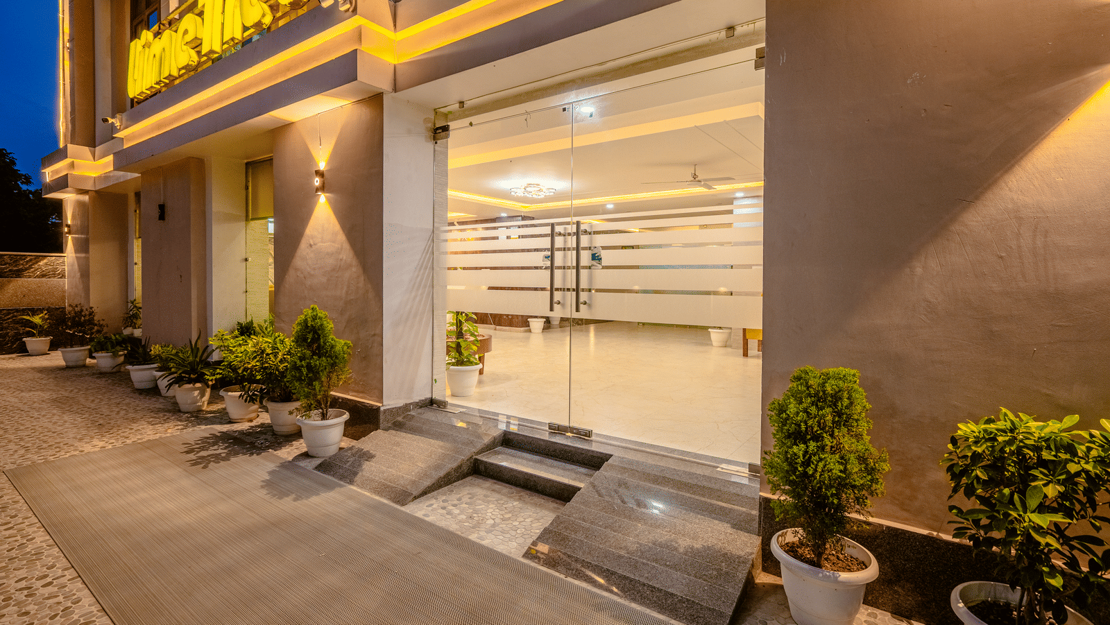 Covered porch entrance of Lime Tree Hotels & Service Apartment Private Limited featuring potted plants and a paved walkway.