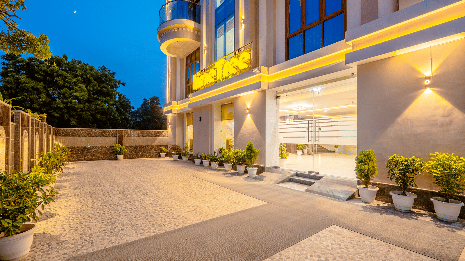 Driveway of Lime Tree Hotels & Service Apartment Private Limited featuring an ornate illuminated facade and potted plants.
