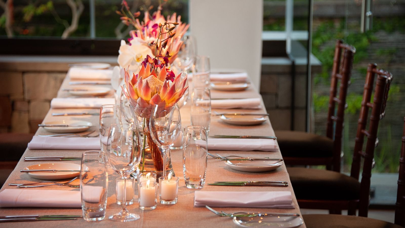 A long dining table adorned with tall orange floral arrangements, candles and a pink table runner, set against a garden view