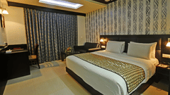 A contemporary room at Pride Ananya Resort Puri featuring a king-size bed with a patterned runner, a seating area with a sofa, a desk, dark patterned wallpaper, and blue curtains.