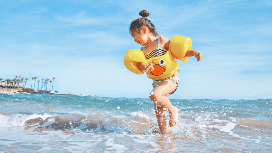 A young child wearing a yellow duck floatie running through shallow beach waves at The Retreat with a bright blue sky palm trees and calm sea visible in the background.