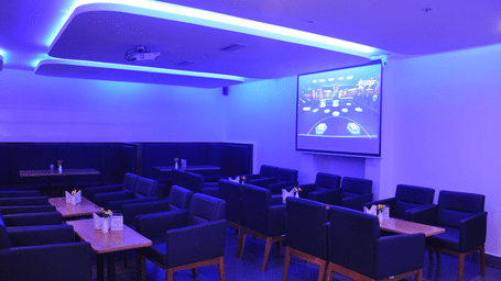 A dimly lit hotel lounge bar featuring rows of seating facing a large projection screen under vibrant blue ambient lighting - Hotel Pandian