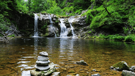 Twin waterfalls cascade down a mossy rock face into a shallow pool with a stacked stone cairn rising from the rocky streambed amid lush green forest