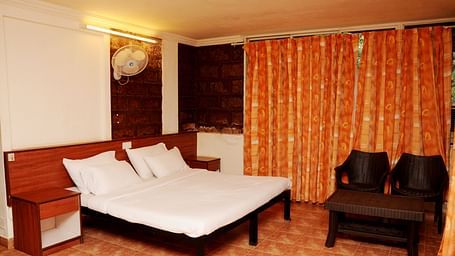 A spacious room at Wind Chalet Resort with a large bed and pillows against a wooden headboard, a bedside table, and a table with a pair of chairs at the foot of the bed, with curtains covering the windows.