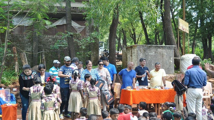 Another large group of people, including children, are gathered outdoors at Forest Escapes Koyna, with a table set up nearby.