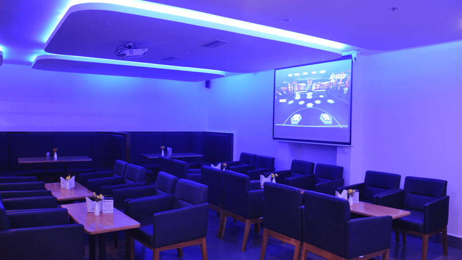 A dimly lit hotel lounge bar featuring rows of seating facing a large projection screen under vibrant blue ambient lighting - Hotel Pandian