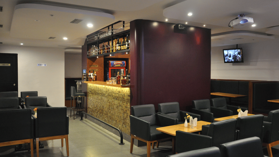 Indoor view of a contemporary hotel bar and restaurant seating area with tables, black leather chairs, and a bar counter - Hotel Pandian
