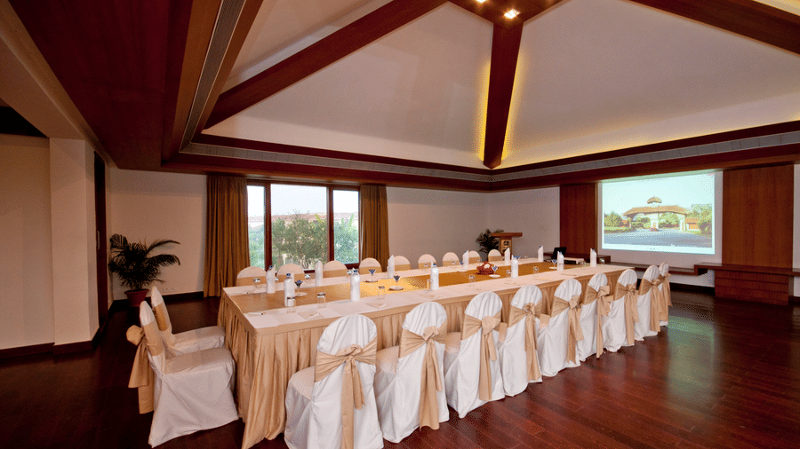 A conference room at Vedic Village Spa Resort featuring a long boardroom table with white chair covers wooden ceiling beams and a projection screen at the far end.