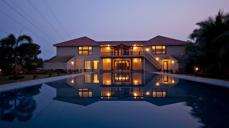 A stunning night view of a resort building at Vedic Village Spa Resort with warm lights reflecting on a calm swimming pool surrounded by palm trees under a twilight sky.