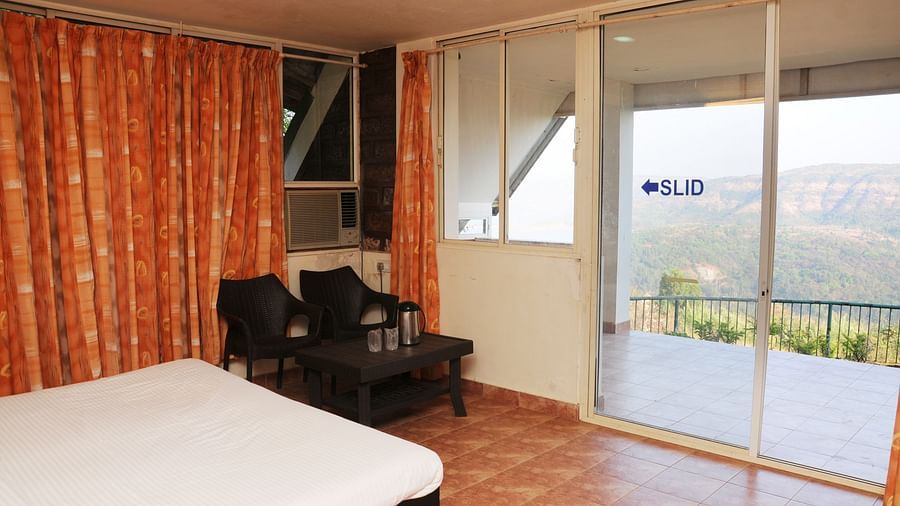 A spacious room at Wind Chalet Resort with curtains covering the windows, a table and chairs placed in the corner, and a balcony offering a view of nature.