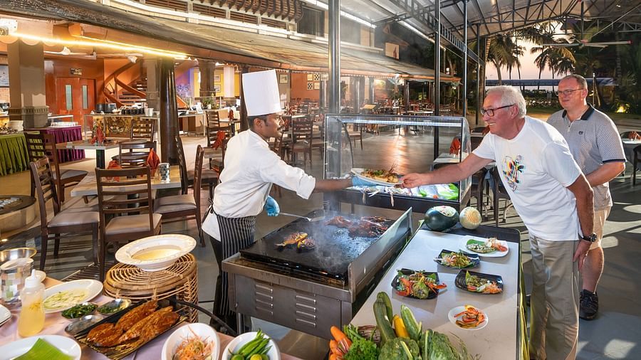 Chef at live grill station serving freshly cooked food to a guest, with another man standing nearby and a display of seafood, vegetables, and salads in a relaxed open-air seaside setting at Uday Samudra Leisure Beach Hotel