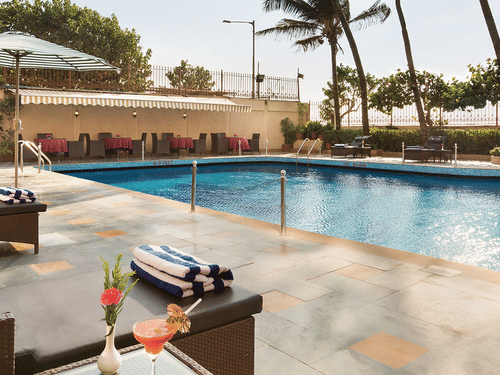 A clear blue swimming pool with drinks in the foreground and surrounded by palm trees at the Ramada Plaza by Wyndham Palm Grove. 