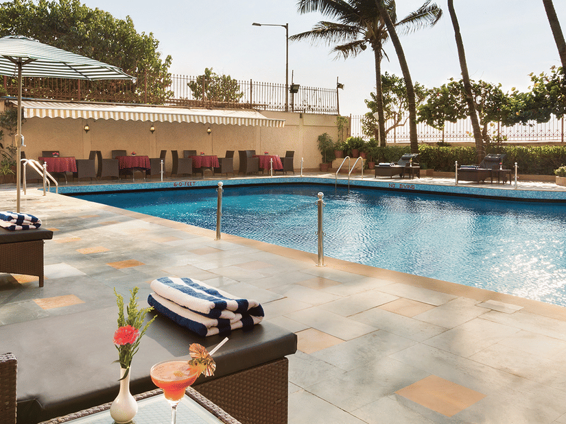 A clear blue swimming pool with drinks in the foreground and surrounded by palm trees at the Ramada Plaza by Wyndham Palm Grove. 