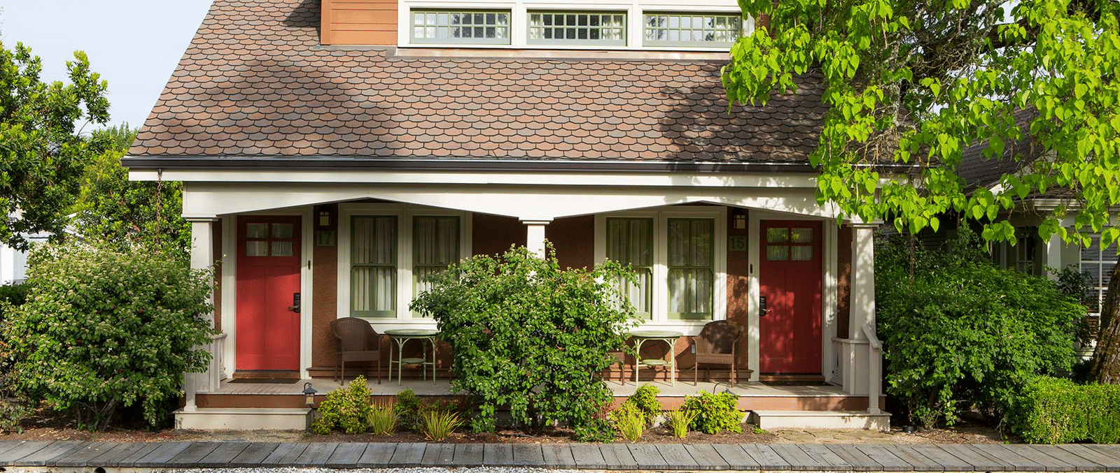A quaint duplex-style house with a covered front porch, 2 matching red doors, outdoor seating, and surrounding greenery viewed from the street at The Tallman Hotel