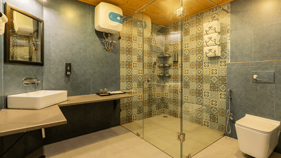 A spacious bathroom featuring a glass enclosed walk-in shower with mosaic tiles, a framed mirror, a wash basin, toiletries, a geyser, and a western toilet | Summit by the Ganges Beach Resort & Spa, Rishikesh